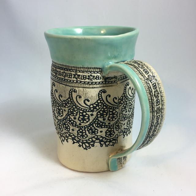 A handmade ceramic mug with aqua accent color at the top and on the handle. An antique lace imprint shows in dark lines on a light surface in the middle.